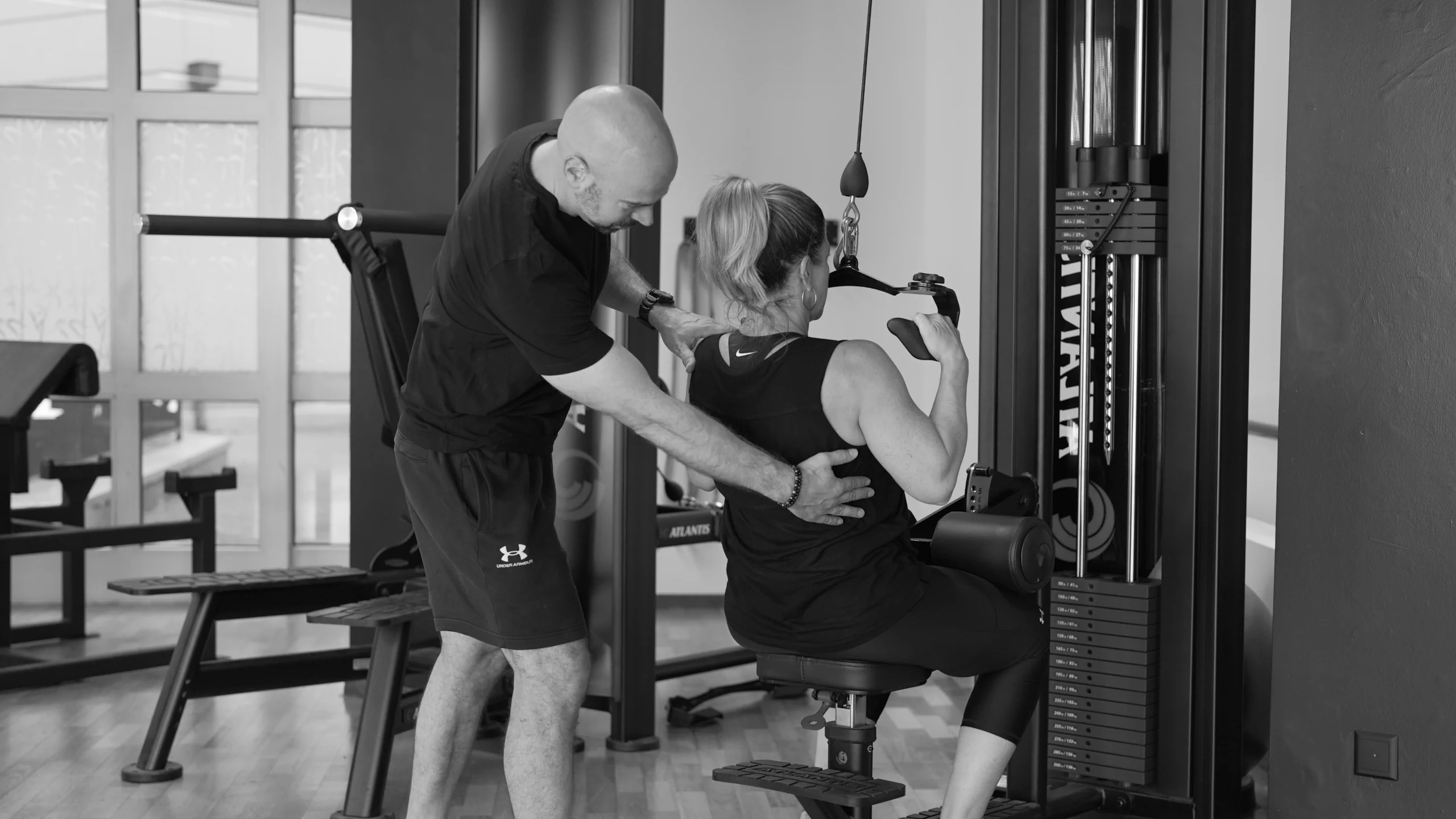 frau-latzug-mit-coach-semper-fi-body-concept-personal-trainer-baden Eine Frau führt den Latzug aus. Der Coach steht hinter ihr und zeigt ihr, welche Muskeln sie trainiert.