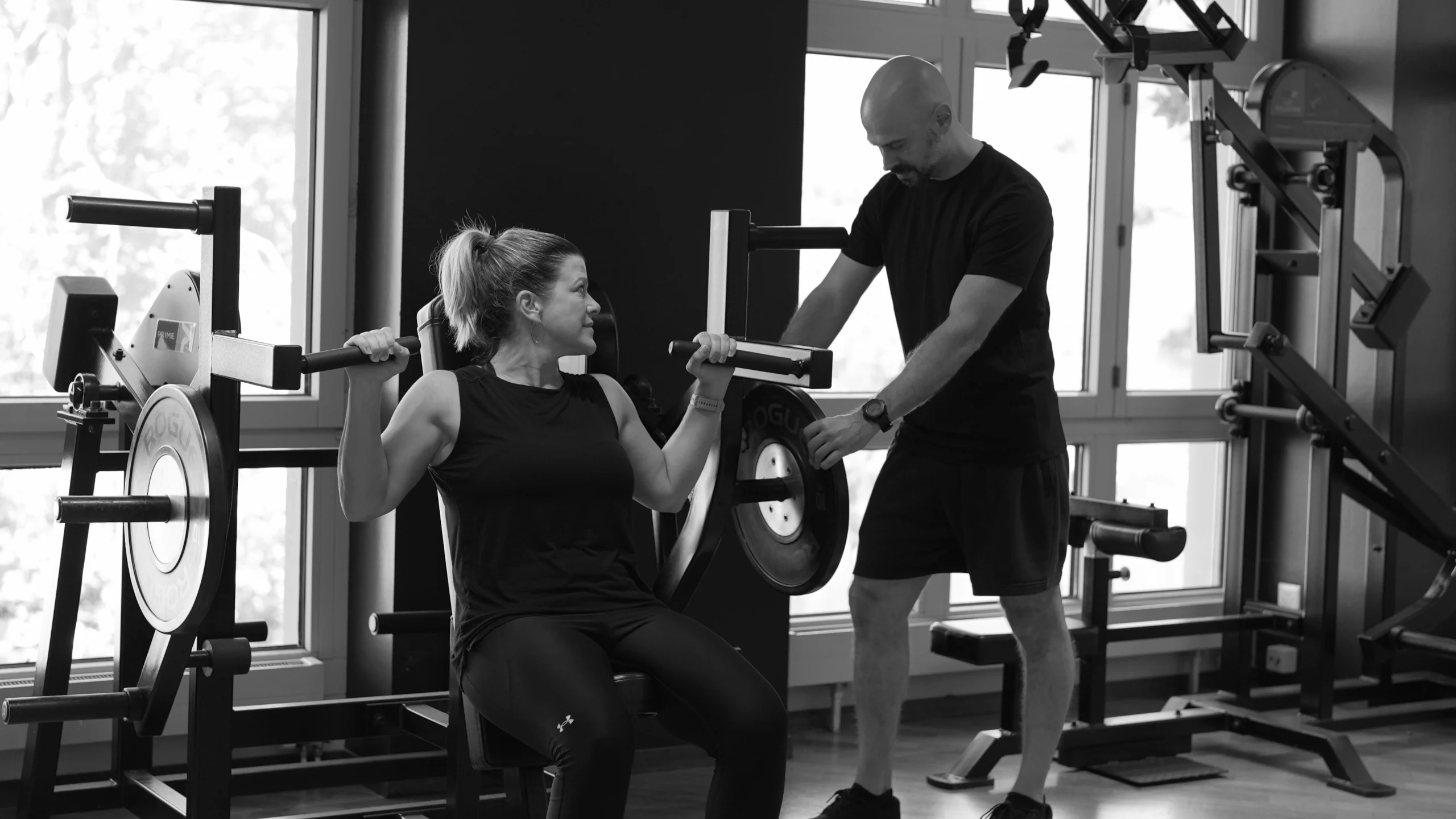 frau-schulterpresse-mit-coach-semper-fi-body-concept-personal-trainer-baden Eine Frau sitz auf der Schulterpresse und schaut zum Coach, während er das Gerät mit Gewichtsscheiben belädt.